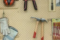 a hammer , pliers , gloves , and screwdrivers are hanging on a peg board .