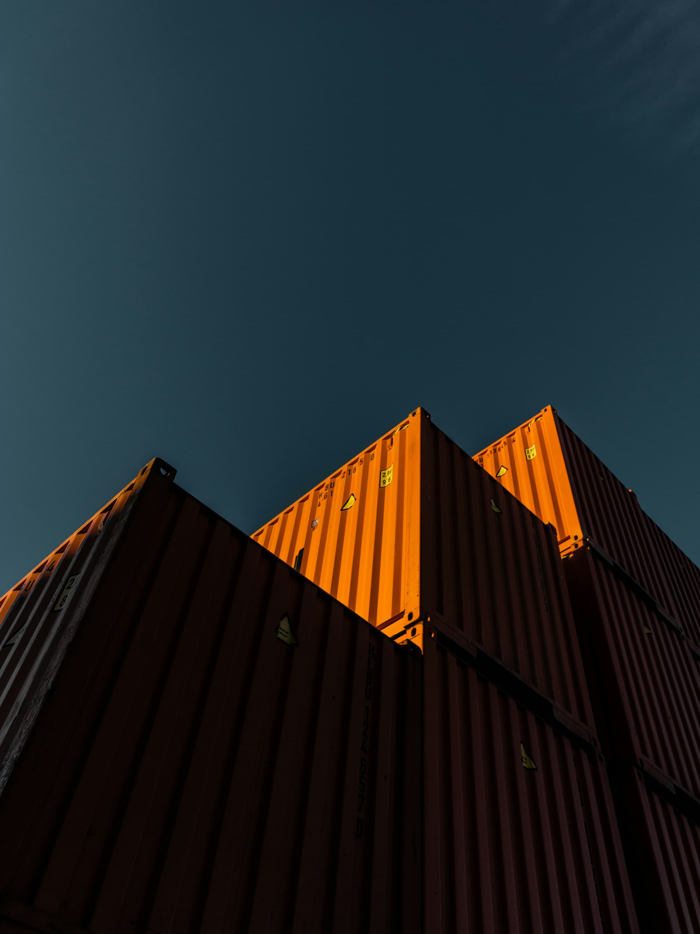 Stacked shipping containers, lit orange by low sun, against a deep blue sky